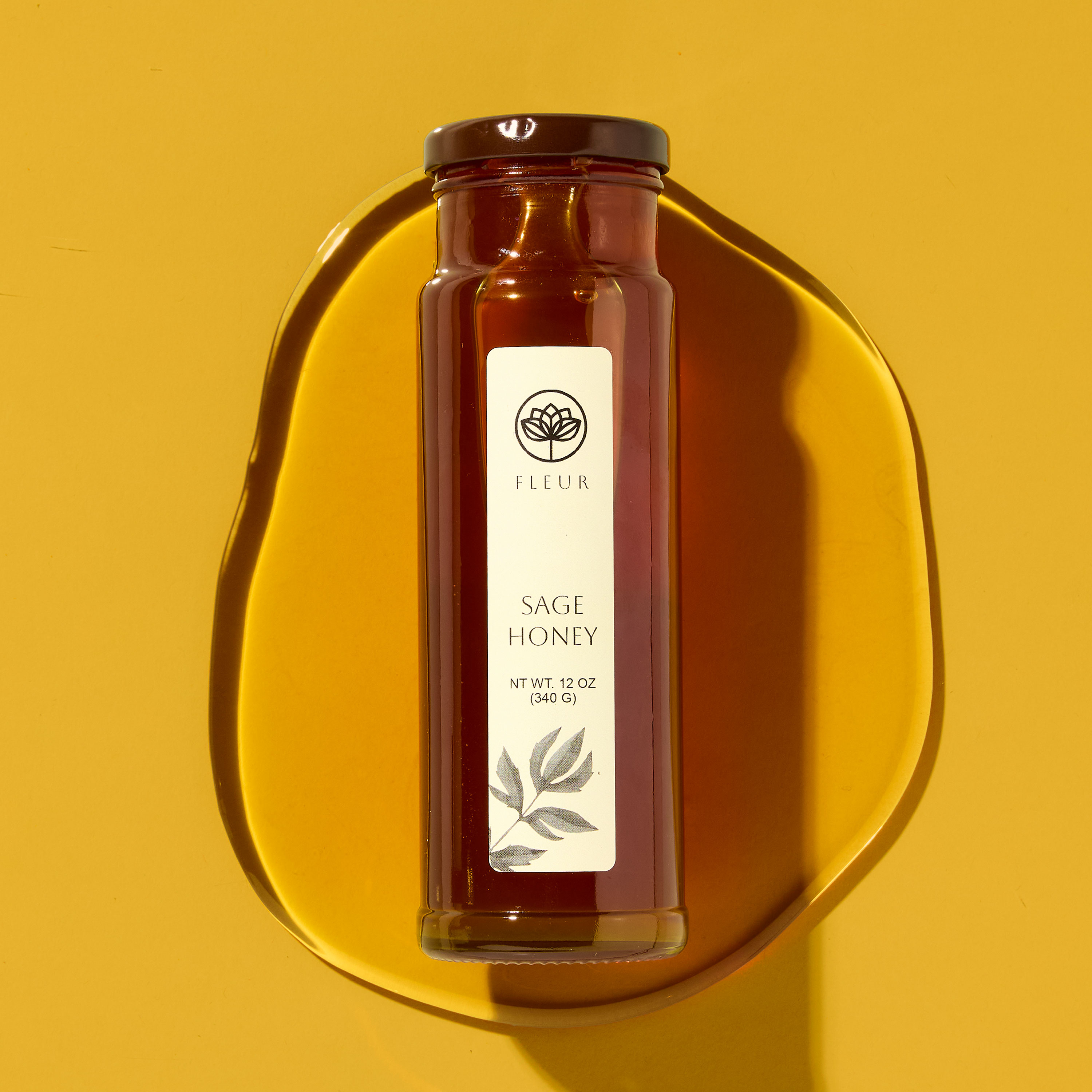 Tall glass jar of sage honey labeled with a minimalist white jar label reading “Fleur – Sage Honey, Net Wt. 12 oz (340 g),” set on a golden yellow background with a pool of spilled honey underneath. The elegant label for jars showcases a soft botanical illustration, perfect for labels for handmade items, or custom labels for jars at a farmers market or boutique food shop.