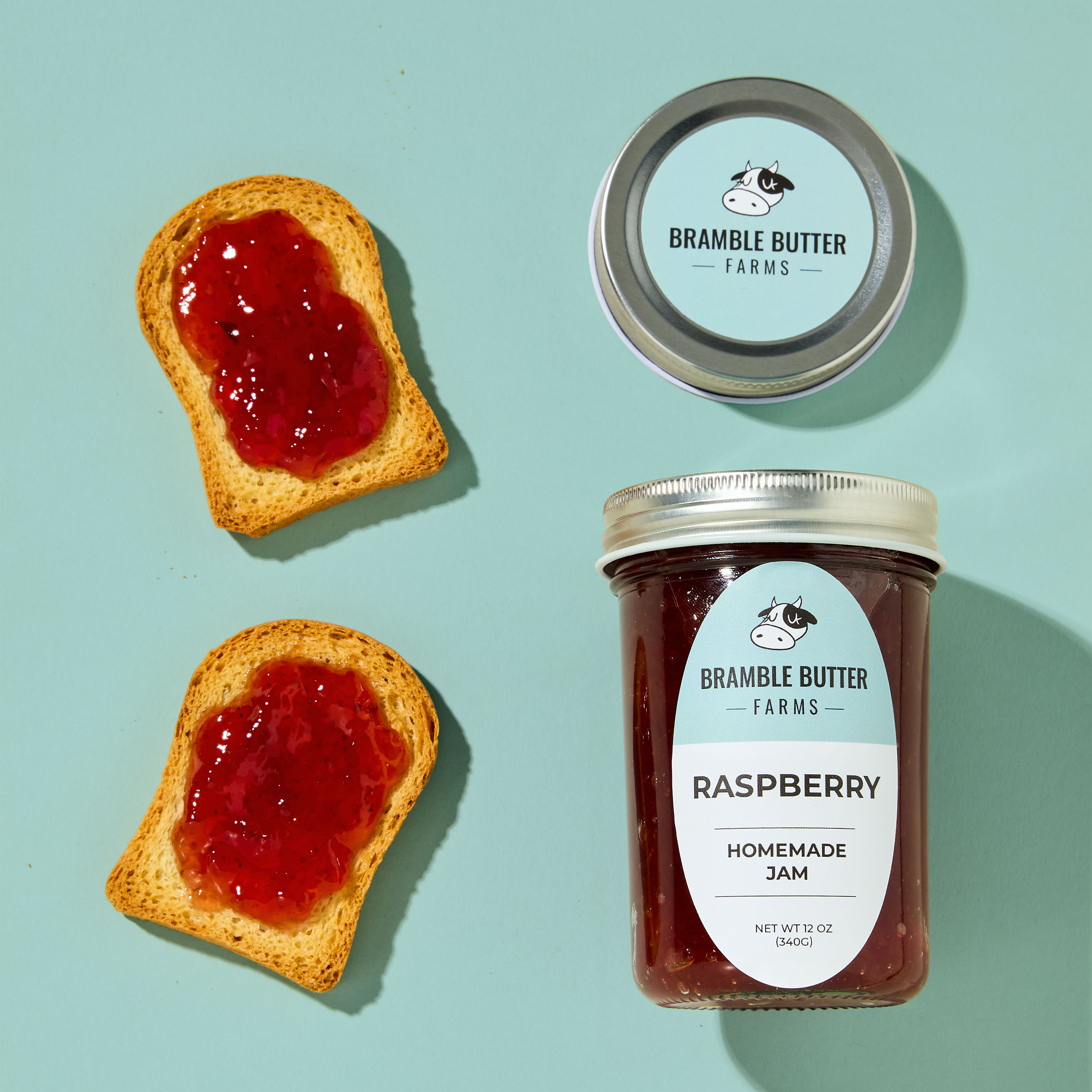 Glass mason jar of Bramble Butter Farms raspberry homemade jam with a pale blue mason jar label and matching round lid label, accompanied by two mini toast slices topped with jam. The clean, oval custom label for jars displays product name, branding, and net weight (12 oz), making it ideal stickers for canning jars and labels for handmade items at markets or gift shops.