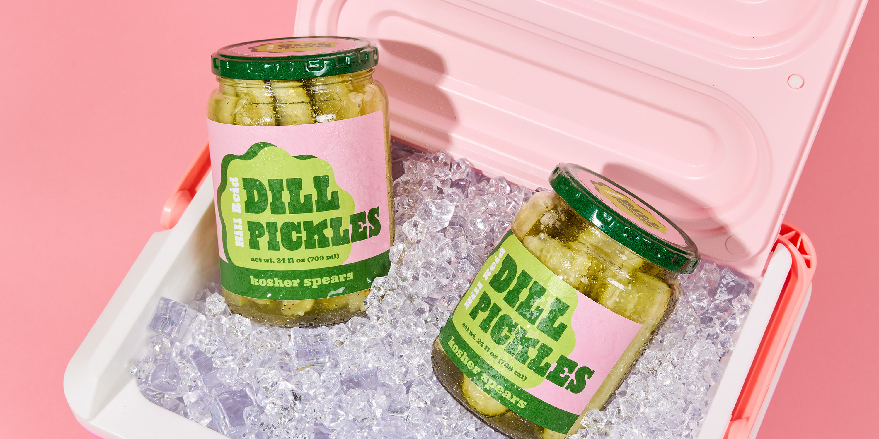 Two glass jars of dill pickles are shown on ice in a pink ice chest on a pink background. The pickle jars are covered in condensation from the ice, but the pink and green labels are fully intact and not smudged.