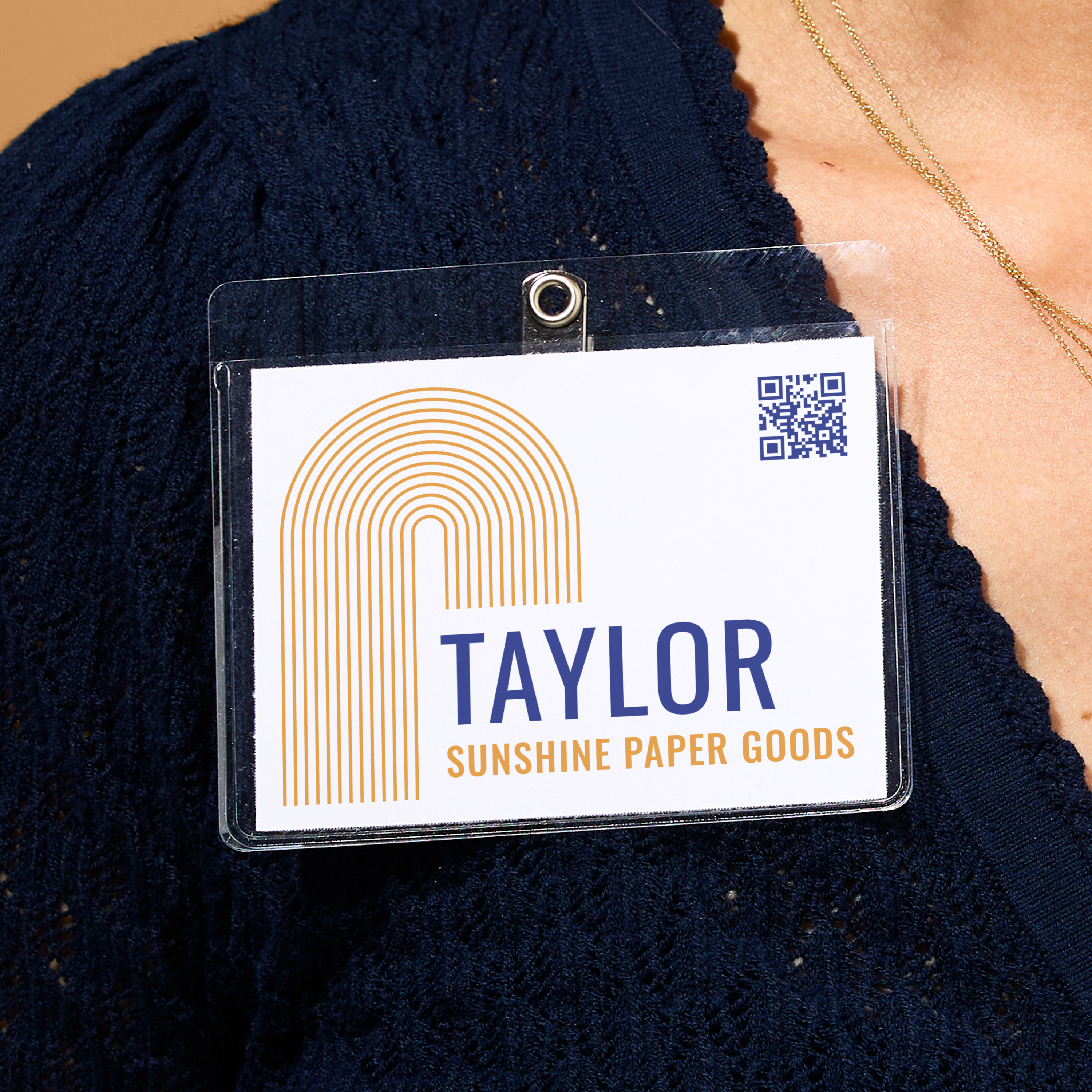 Close-up of a business name tag for Sunshine Paper Goods, clipped to a navy blue blouse. The badge shows a modern gold arch logo, bold blue text reading "TAYLOR," orange subtext, and a small blue QR code in the corner.