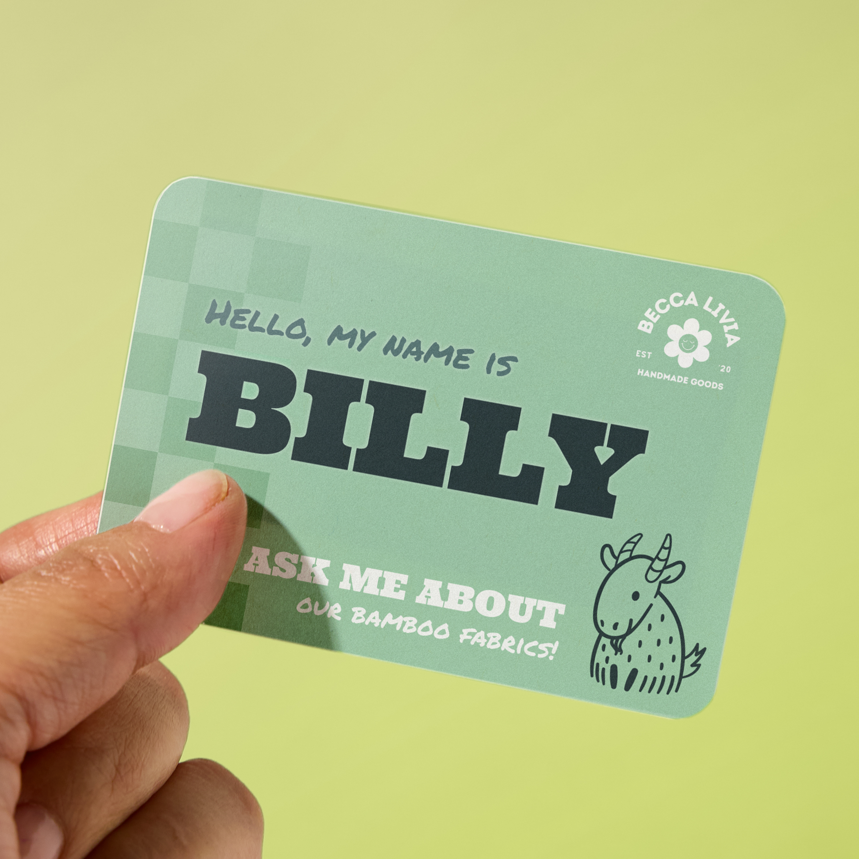 Green “Ask Me About” name tag design for trade shows, held in hand against a light green background. Text reads “Hello, My Name Is Billy” with bold black lettering and a cartoon goat illustration promoting bamboo fabrics for Becca Livia Handmade Goods.