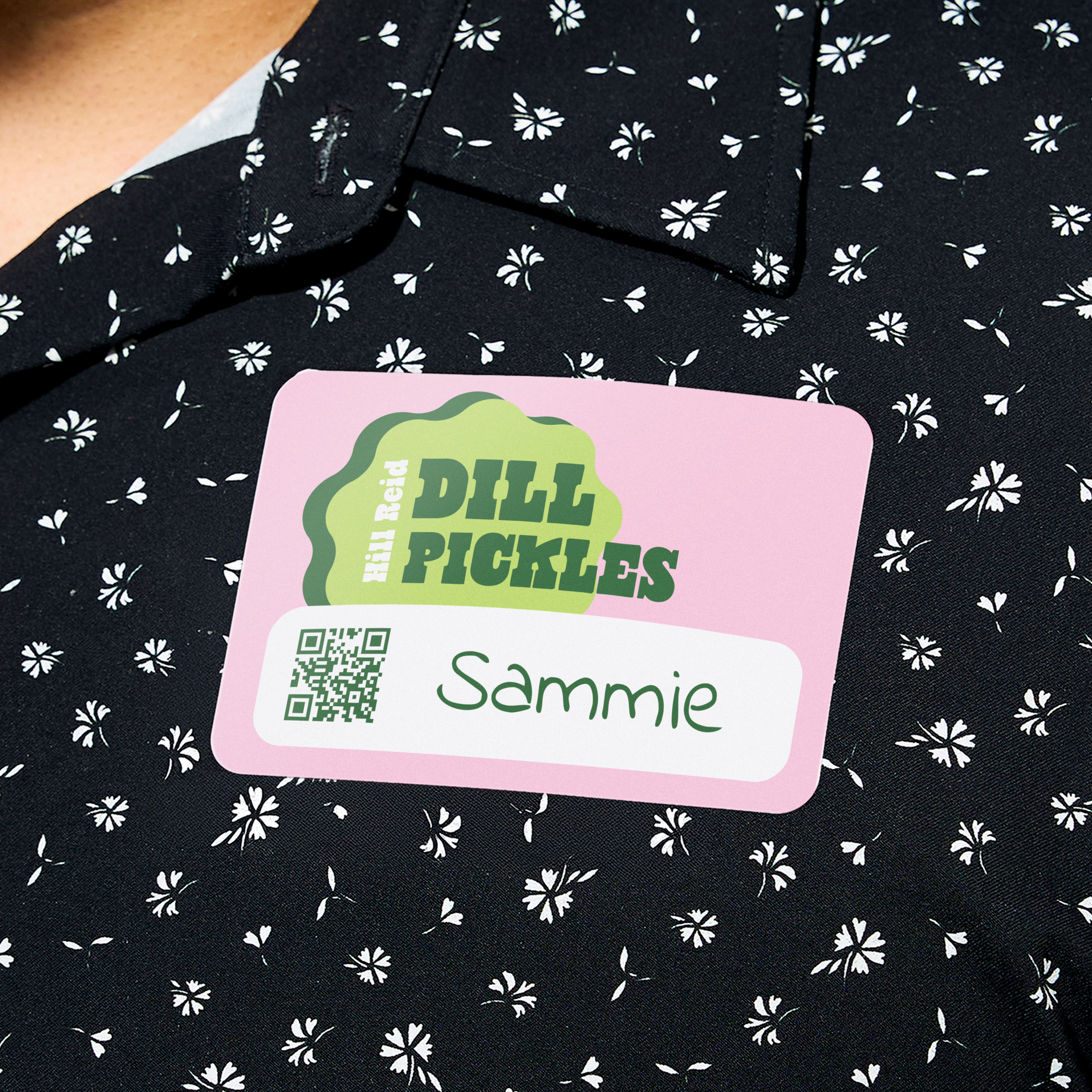 Networking name tag idea with QR code for business events. Pink name badge reads “Dill Pickles” with green logo and “Sammie” in script font. Attached to a black floral shirt.