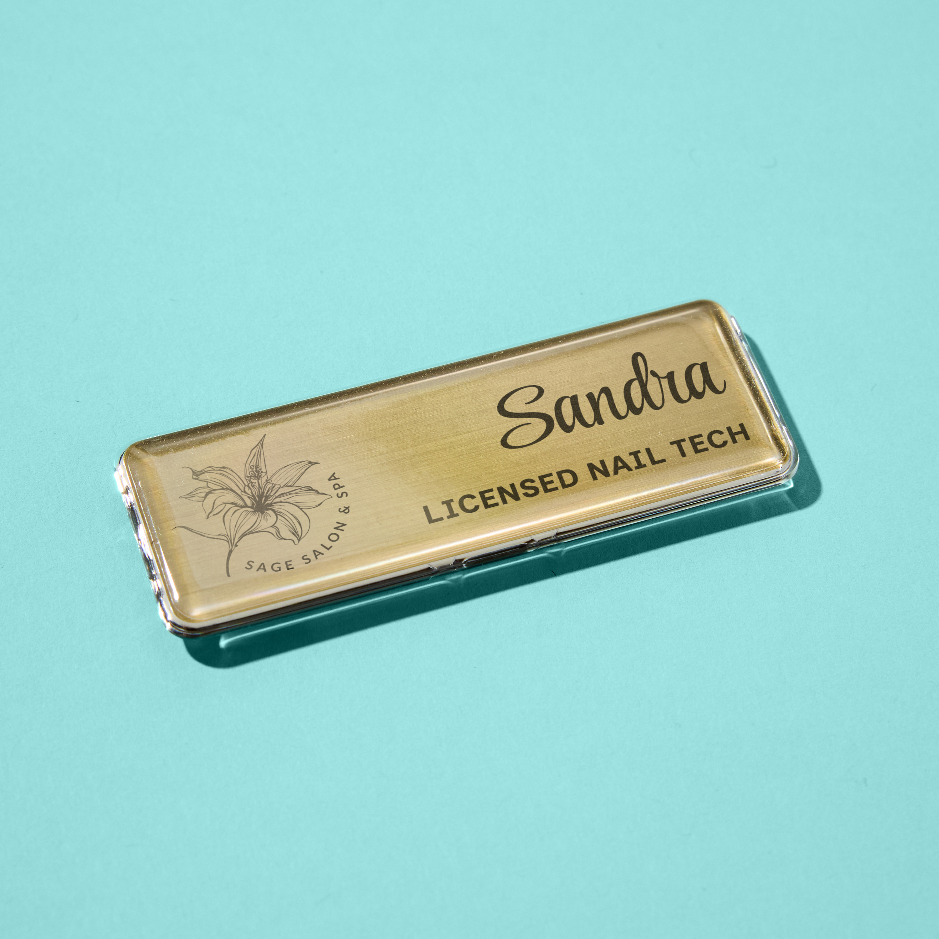 Professional metal name badge for client-facing staff. Gold rectangular Mighty Badge displays “Sandra, Licensed Nail Tech” with a floral Sage Salon & Spa logo. Shown on aqua background.