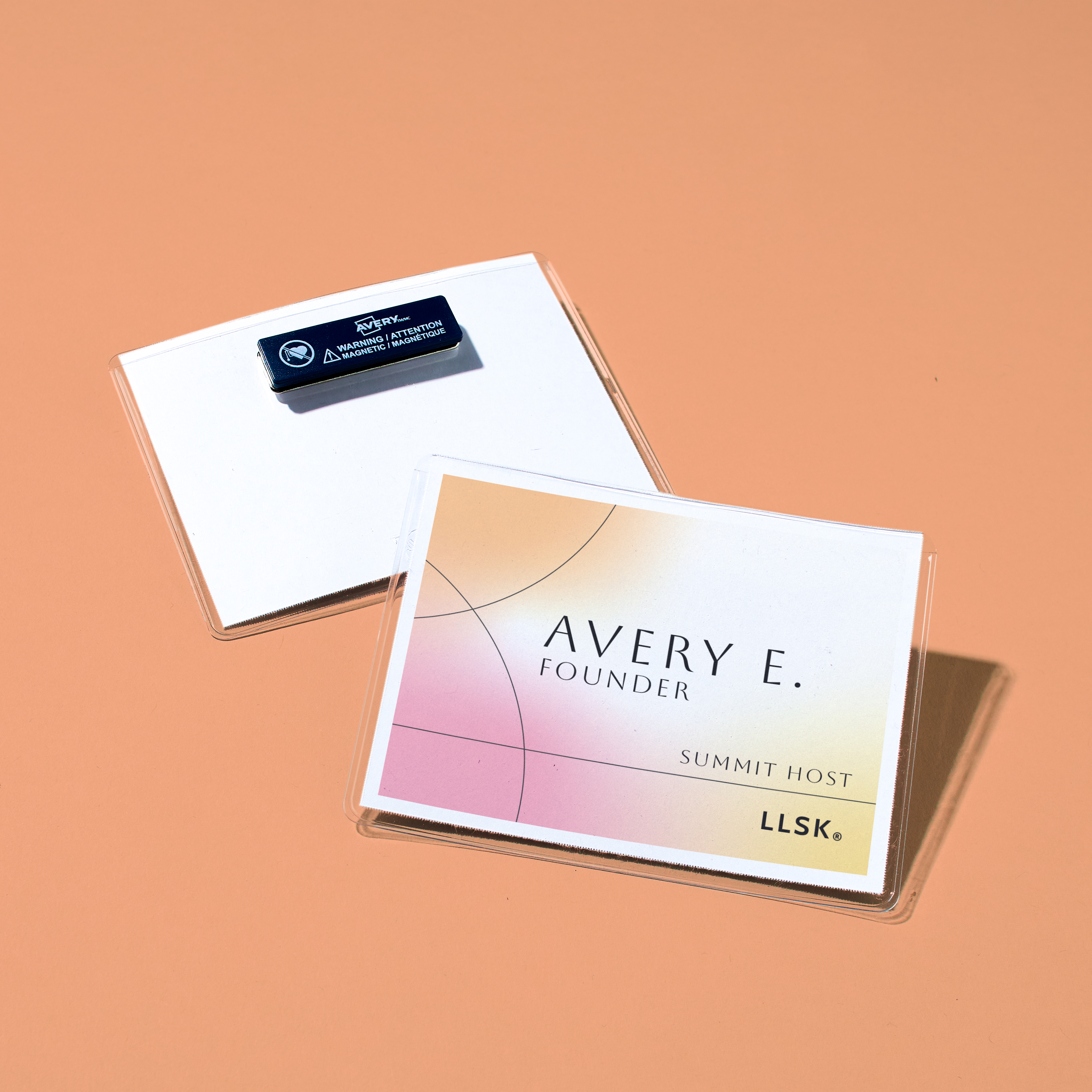 Two rectangular name badges rest on a peach-colored surface. The front badge features a sleek, modern design with soft gradient tones of peach, pink, and gold. It displays the name “Avery E.” with the title “Founder” and the role “Summit Host” in smaller text. The lower right corner includes the acronym “LLSK.” The second badge is flipped to show a magnet-style backing with the label “Caution: Magnetic Device. Do Not Use with Pacemaker.”