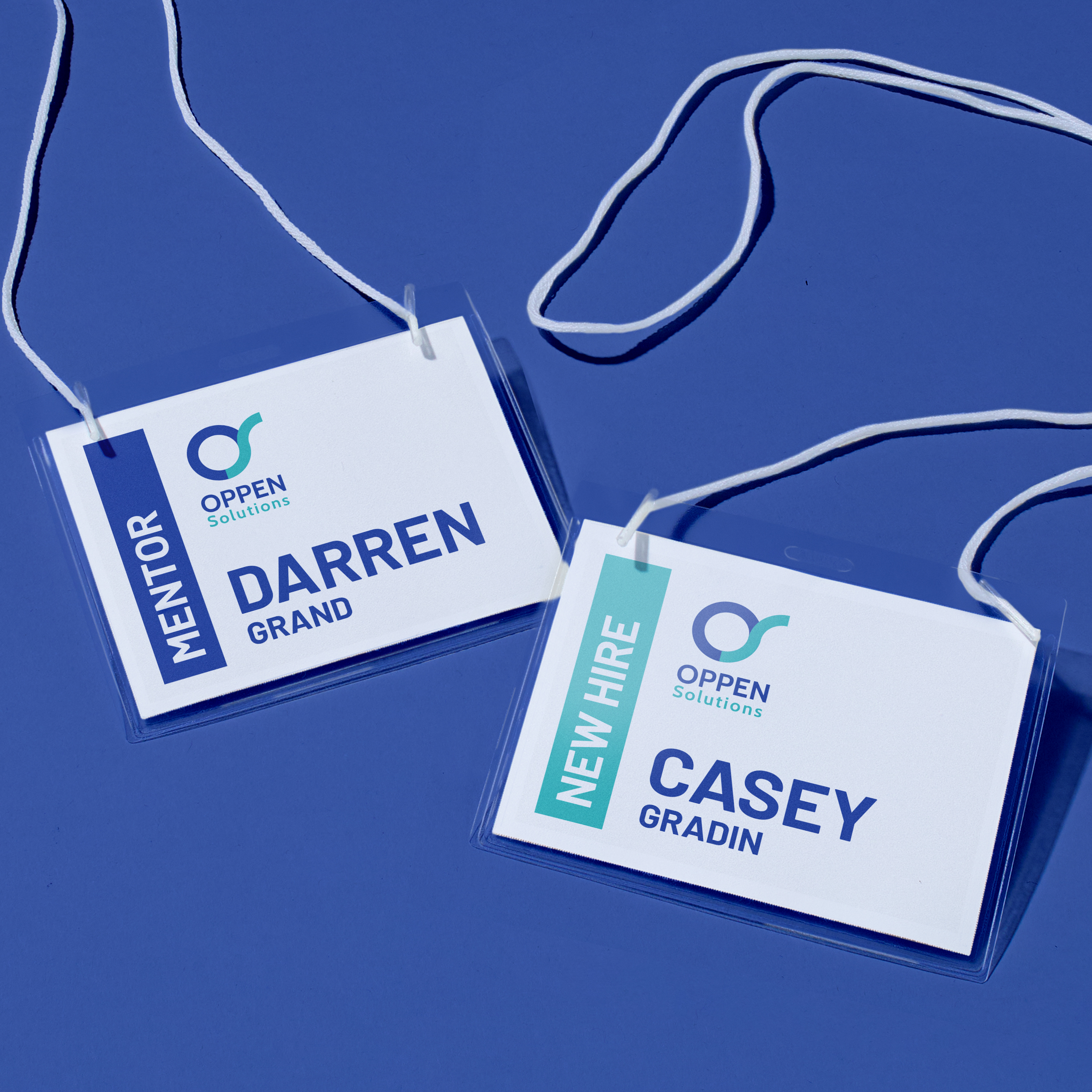 Two horizontal name badges with white string lanyards lie on a cobalt blue surface. The badges display the logo for Oppen Solutions and are labeled with roles and names. The left badge reads “MENTOR” in a vertical blue bar, with the name “Darren Grand” underneath. The right badge reads “NEW HIRE” in a teal vertical bar, with the name “Casey Gradin” underneath. Both badges feature a clean, modern design with bold typography and color accents for easy role recognition.