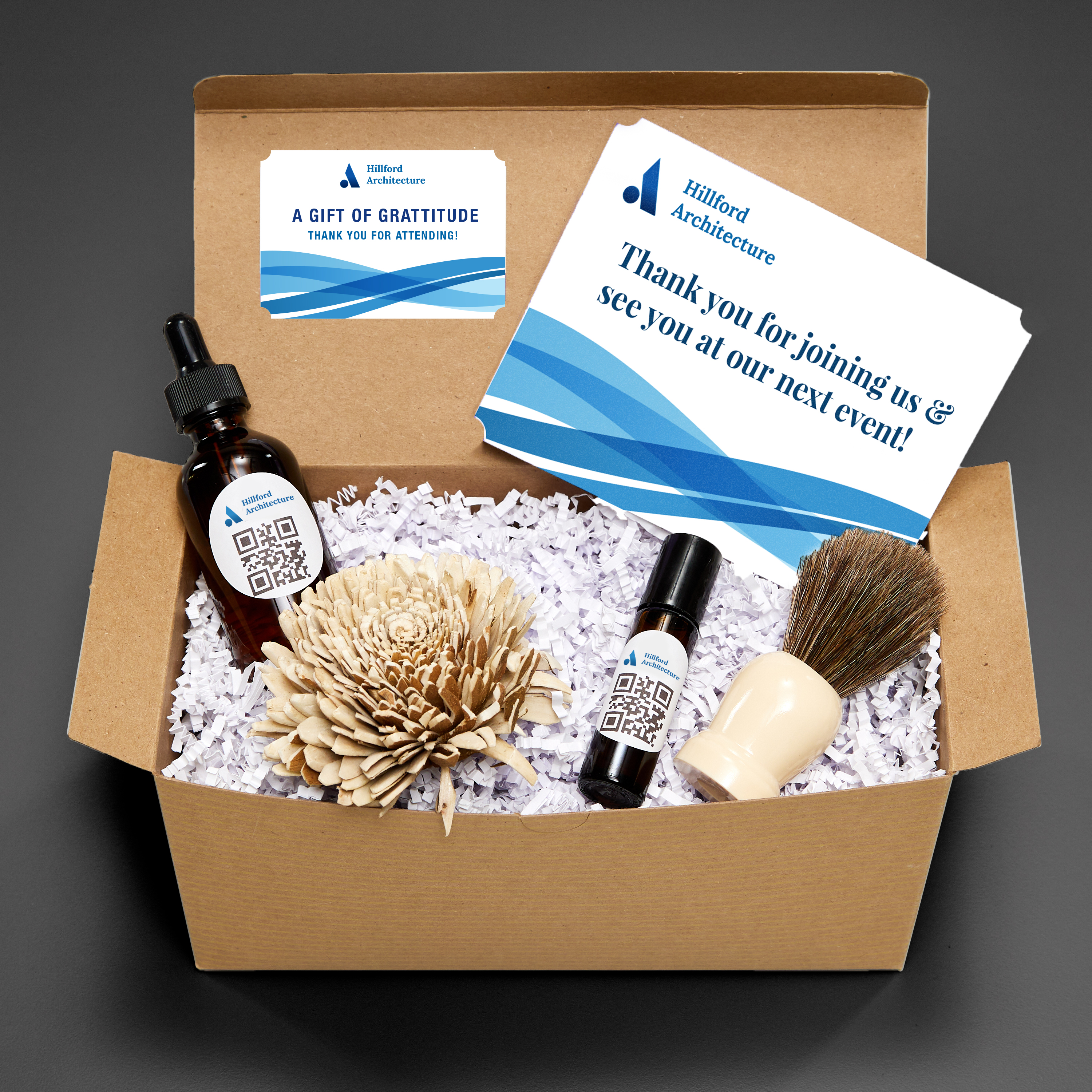 Swag bag idea for bundling products with like items. Glass apothecary bottles are shown in a cardboard box with a shaving brush and a custom-printed postcard. 